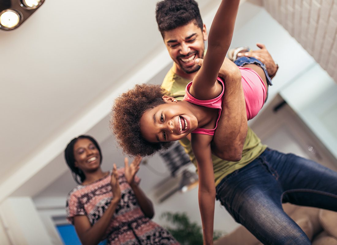 About Our Agency - Happy Father Plays With His Daughter at Home as the Mother Watches and Smile at Home