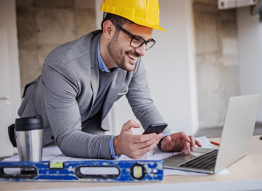 Insurance by Industry - Smiling Construction Engineer Using a Laptop and Smart Phone at a Project Site